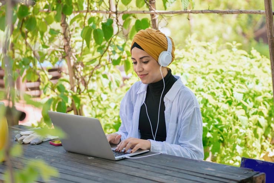 Eine junge Frau mit Turban und Kopfhörern sitzt an einem Laptop in einem sonnigen Garten und sichtet Ihre Altersvorsorge in der Digitalen Rentenübersicht.