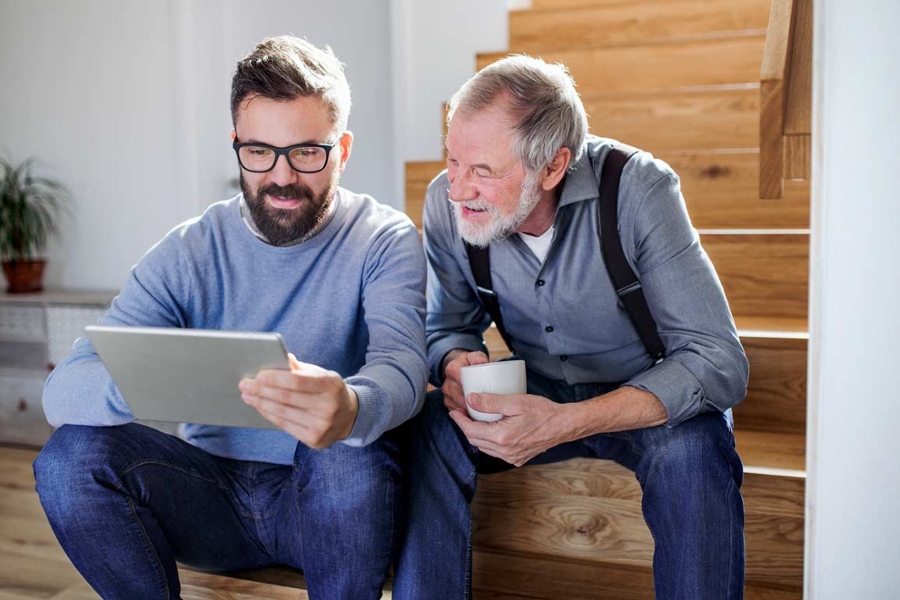 Ein Mann sitzt mit seinem Vater auf einer Holztreppe im eigenen Heim. Auf einem Tablet zeigt er ihm die Digitale Rentenübersicht.
